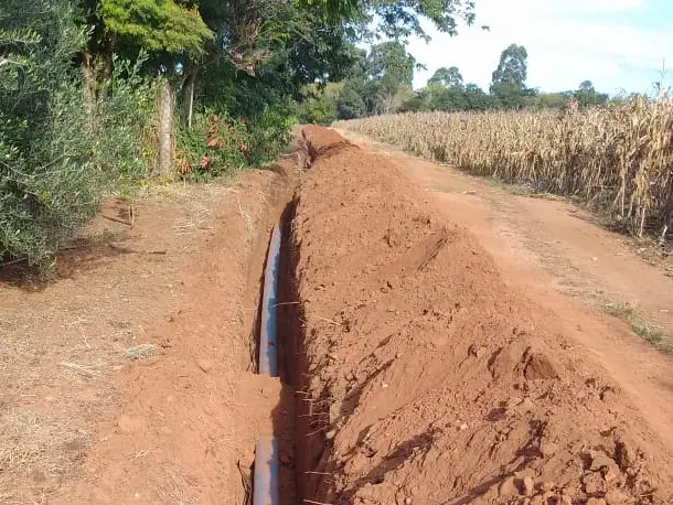 Vala aberta em terra vermelha com tubulação instalada, ao lado de estrada de terra